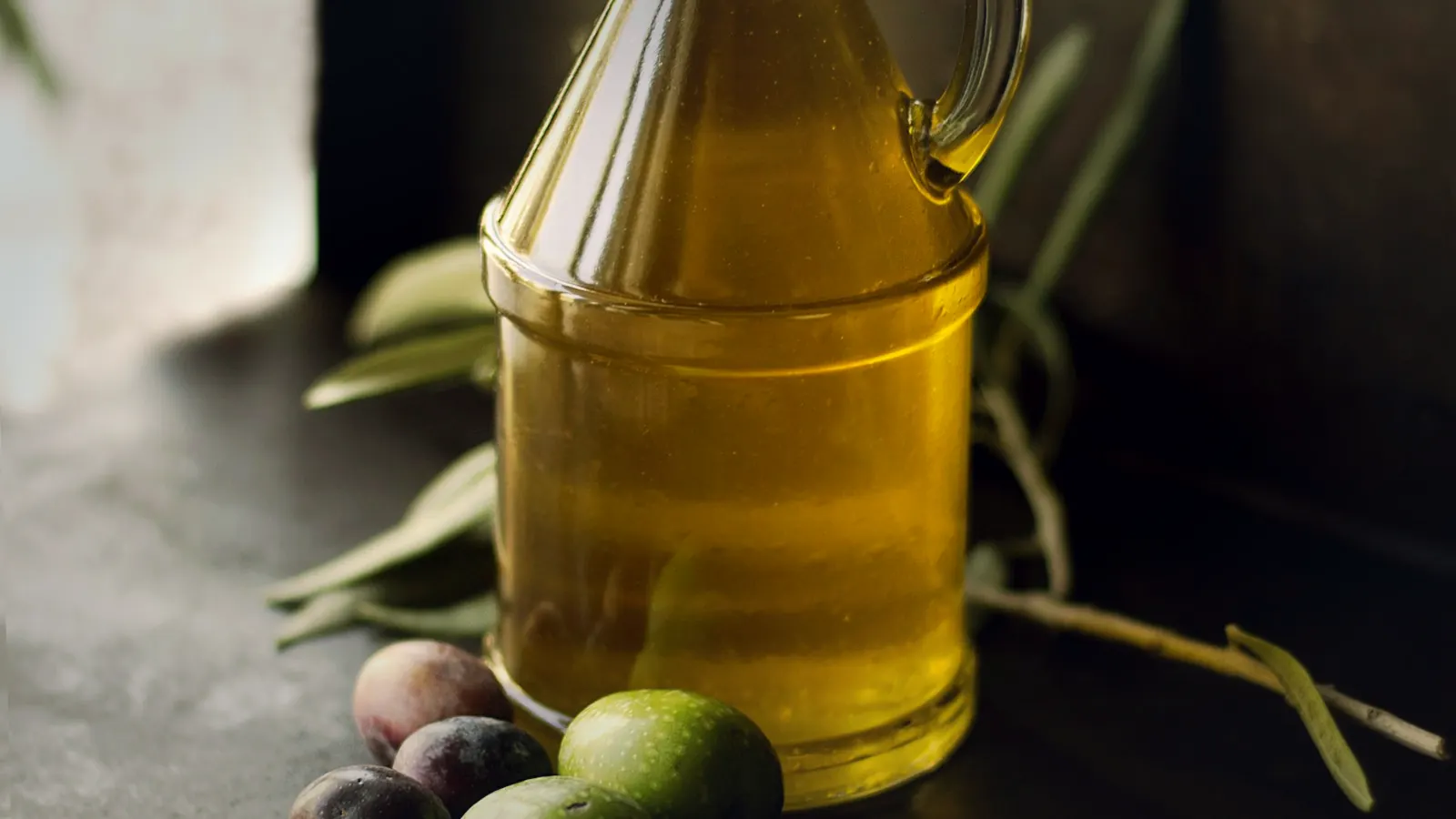 Glass bottle of golden olive oil with scattered green and dark olives on a dark table in warm editorial light