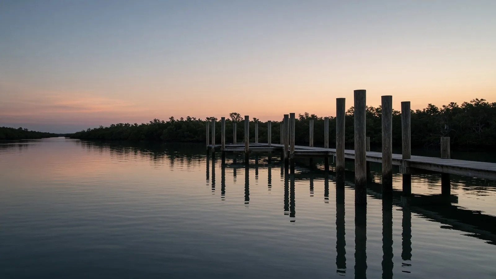 Quiet Gulf Coast channel at soft pre-dawn light with wooden dock pilings, calm reflective water and distant mangrove shoreline