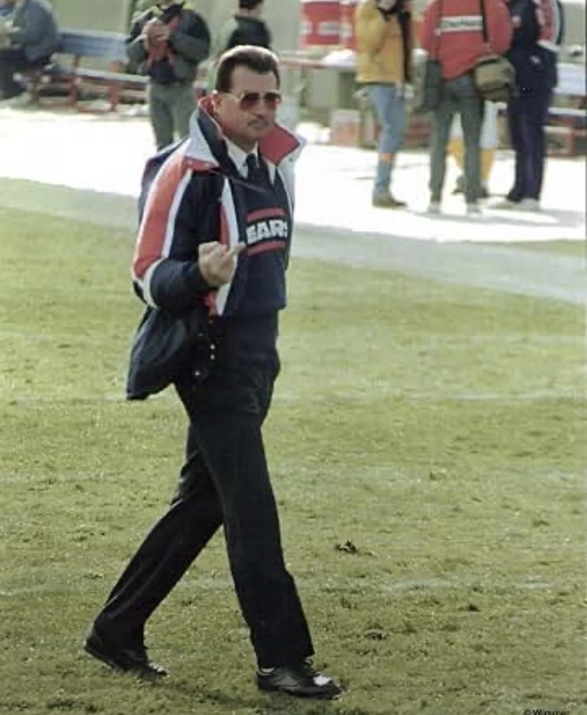 Mike Ditka in a Bears jacket and tinted glasses walking the sideline, giving his trademark one-finger salute